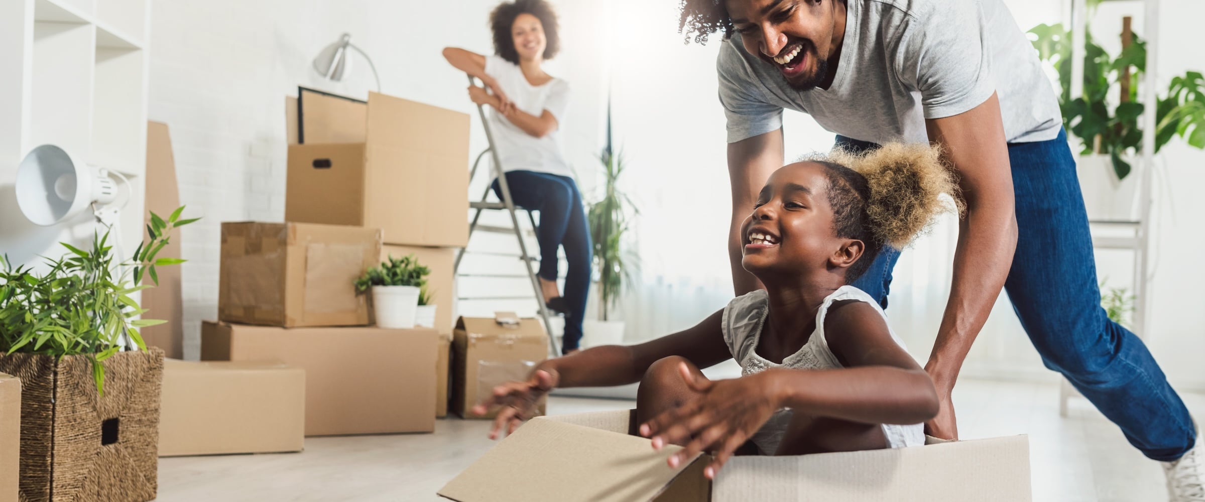 Photo of African-American family in new home. Dad is pushing daughter in cardboard moving box.