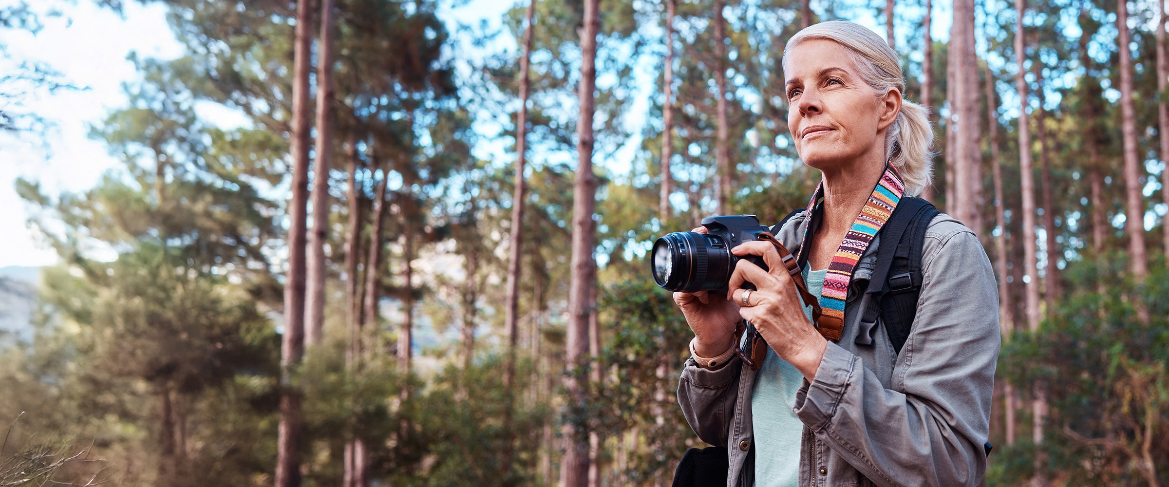 Photo of older woman in woods holding camera and ready to take a picture.