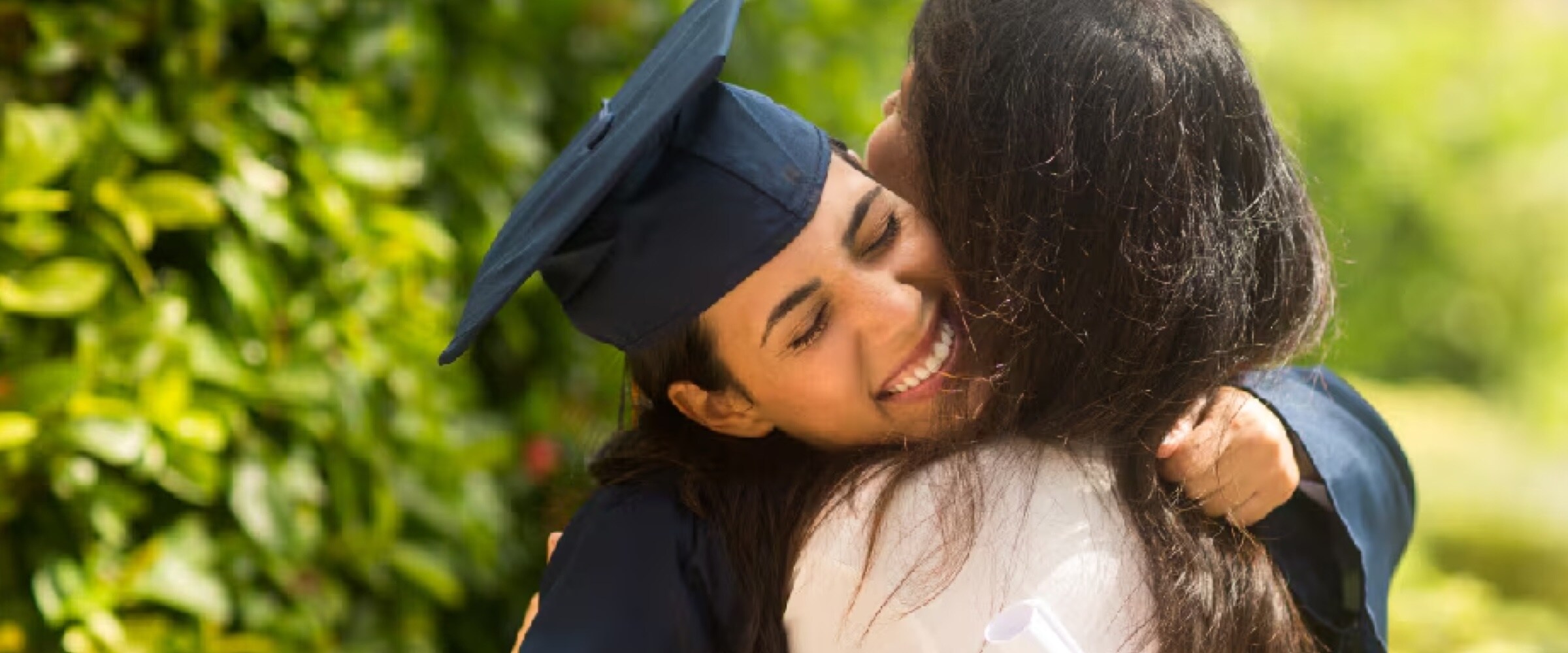 Photo of young woman in graduation gown hugging an older woman.