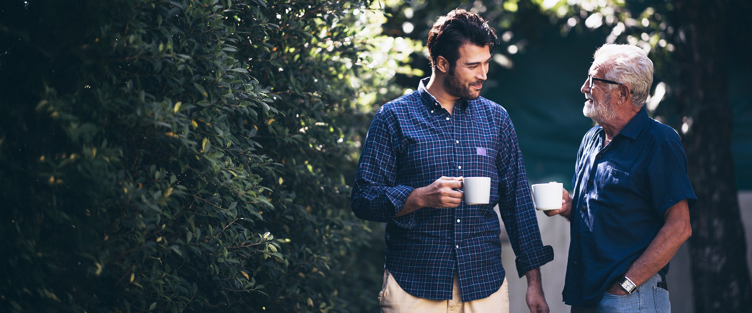 Son and older dad holding coffee mugs and talking outside.