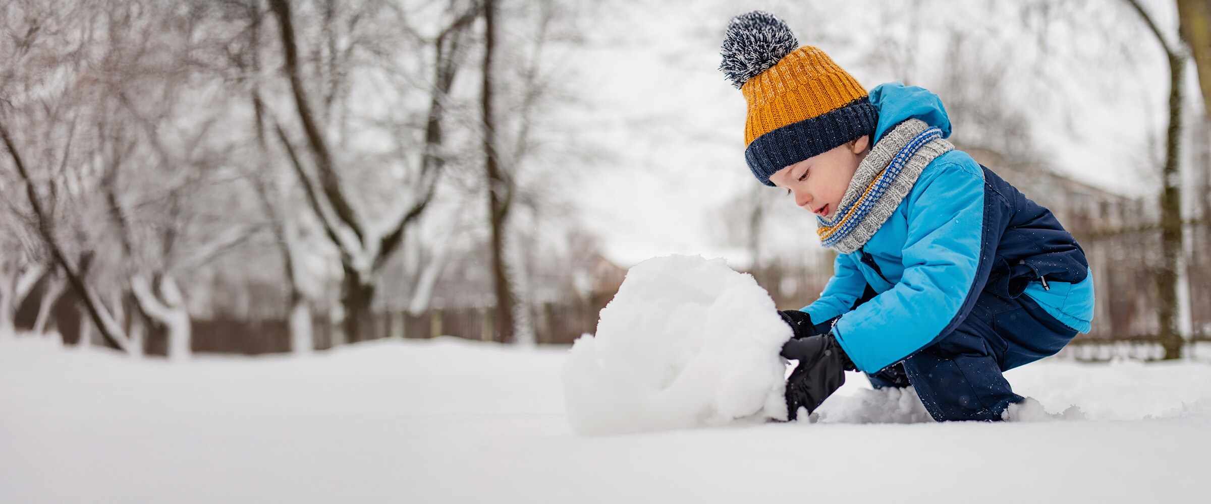 Photo on young child in snow gear rolling a snowball in the snow.