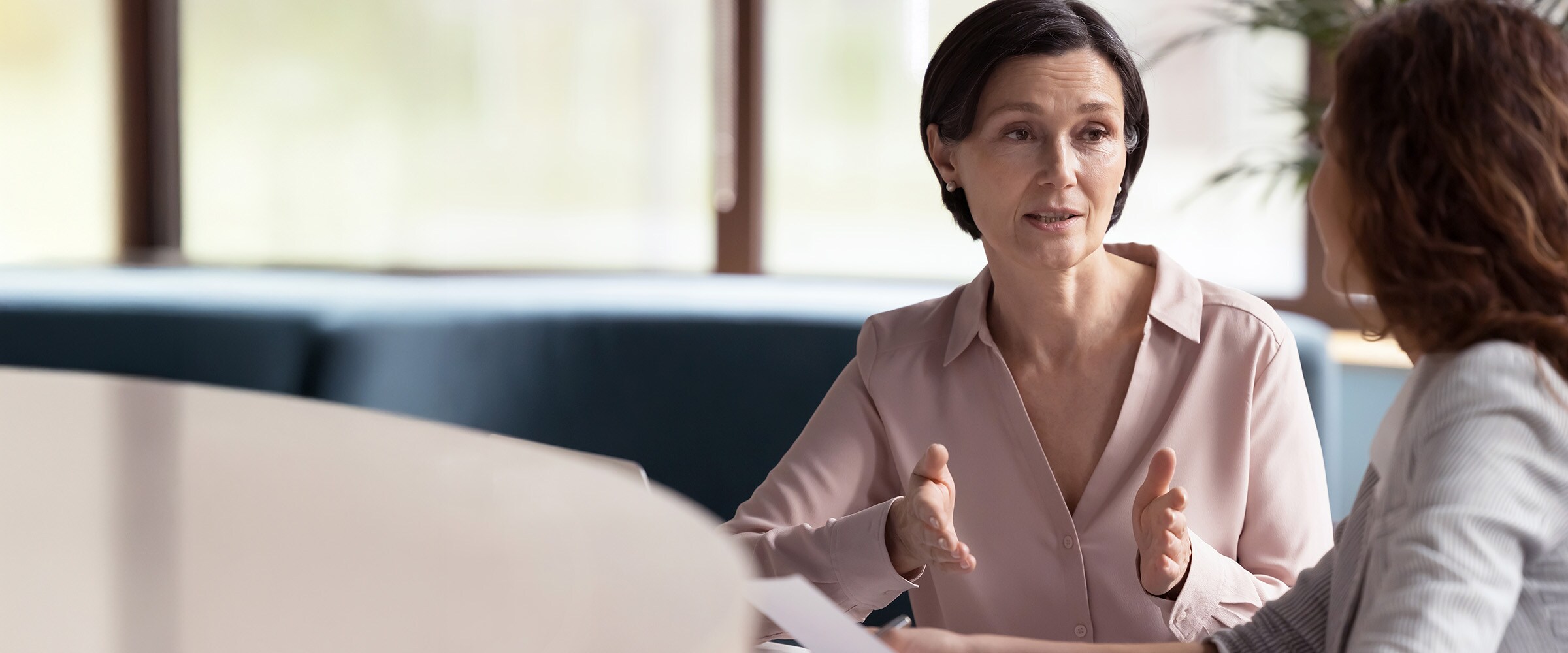 Photo of woman talking to another woman. Both are sitting around a long meeting table.