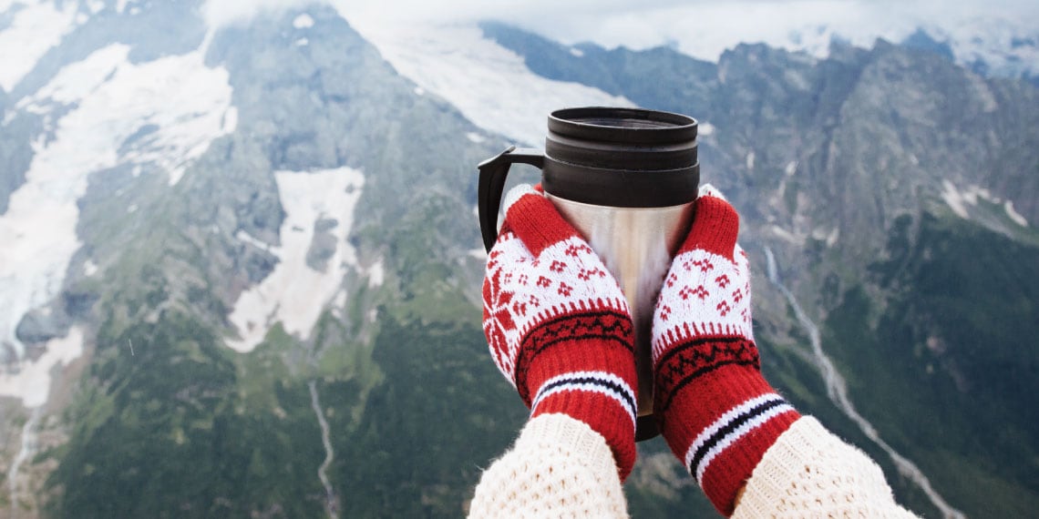 Photo of hands holding a tumbler with view of wintery mountains in the background.